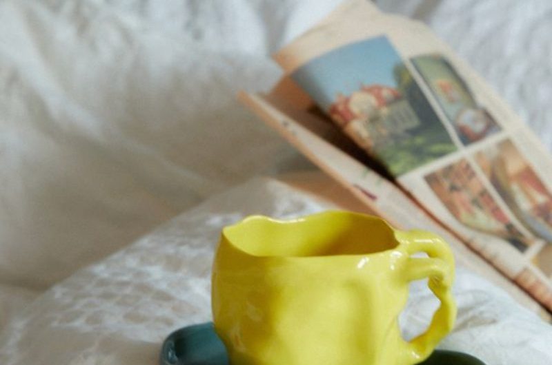 A yellow cup on a green saucer on a bed with white linen and a newspaper.