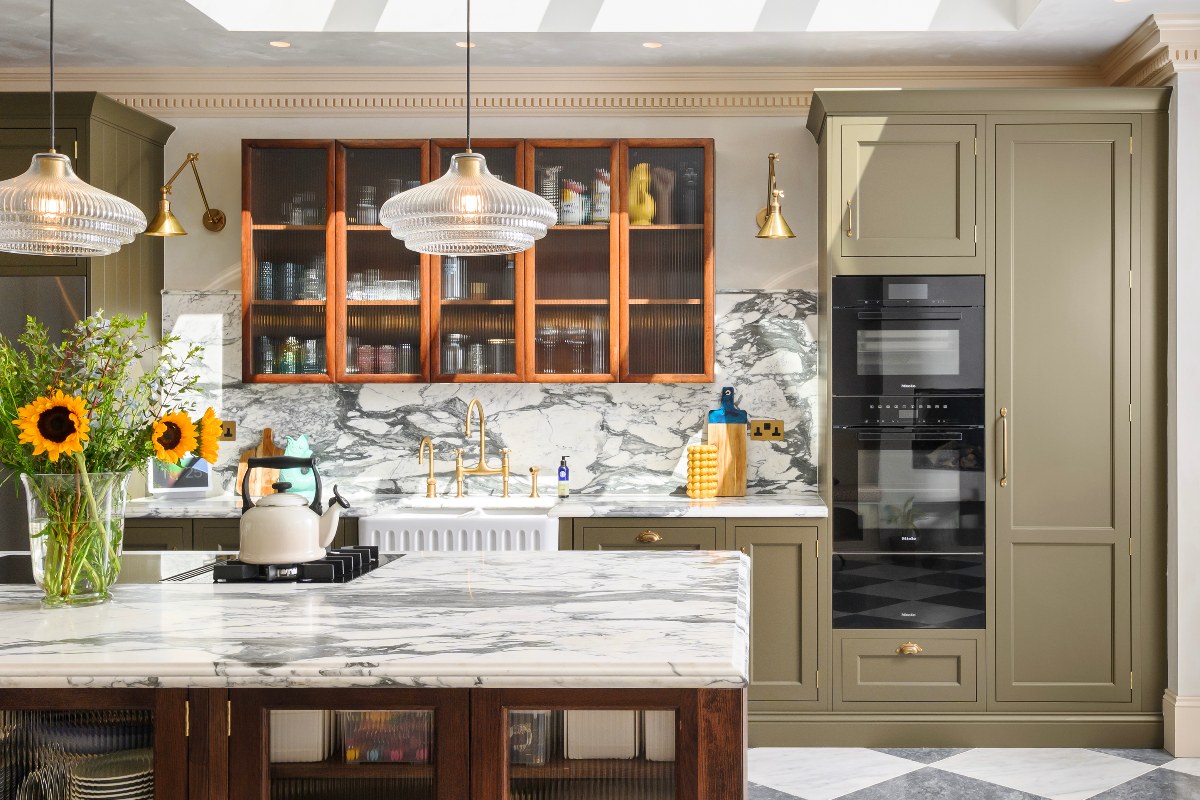 View of the farmhouse kitchen with olive green run of cabinets, built-in appliances, a wood wall-mounted cupboard with fluted glass, a sink zone with marble splashback and a central wood island with matching marble surfaces and a hob.