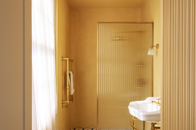 A yellow bathroom design with a walk-in shower, a reeded panel, gold brassware, a classic white basin on a brass frame, and patterned flooring.