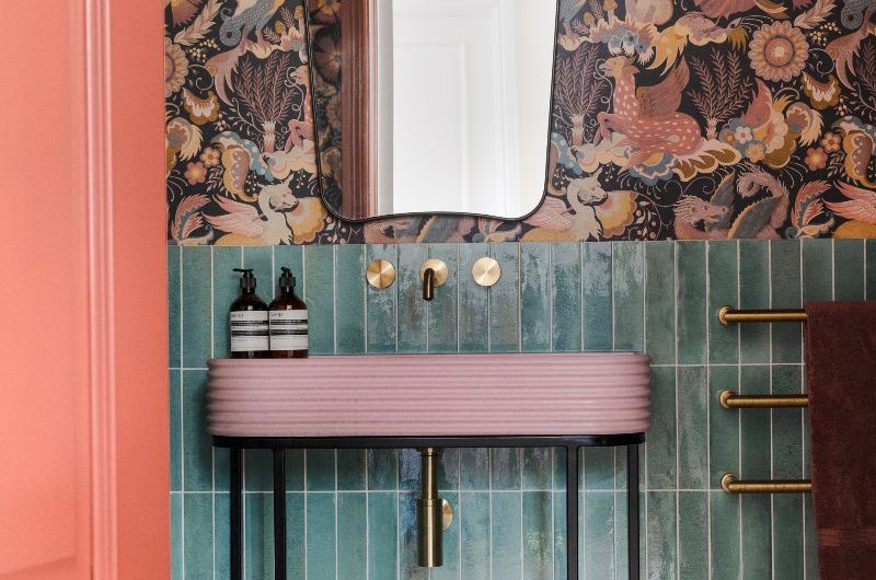 A colourful downstairs bathroom design with green metro tiles on the bottom half of the wall, patterned wallpaper on the upper half, a mirror, wall light and a pink concrete basin on a black frame as you open the door.