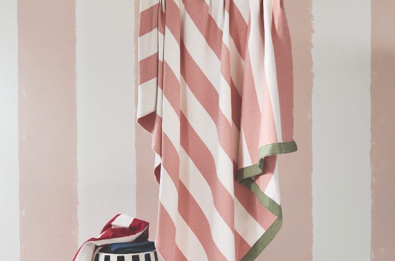A hanging striped throw in pink and sage green with striped wallpaper in the background and a black and white striped stool next to it.