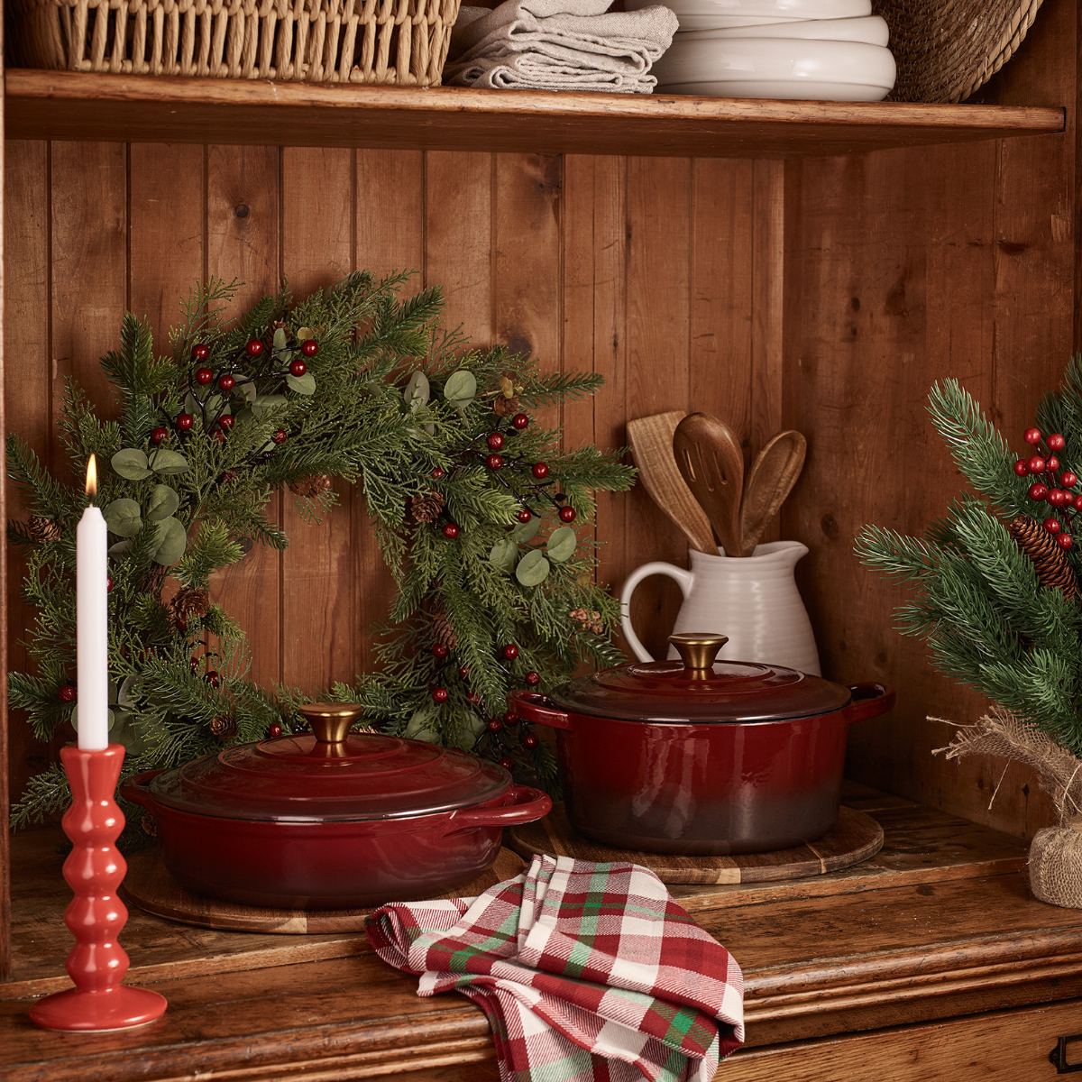 A wood kitchen dresser with red cast iron pots, wreath decorations, and open shelving.