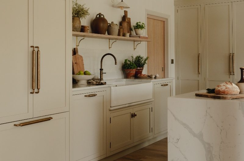 Closeup at the sink zone with a butler sink, marbled surfaces, brass tap, and open wood shelf, and wall lights.