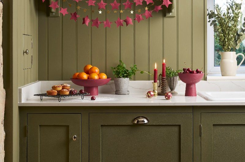 An olive green kitchen design with panelling, classic cabinetry, and festive Christmas decorations in burgundy shades.
