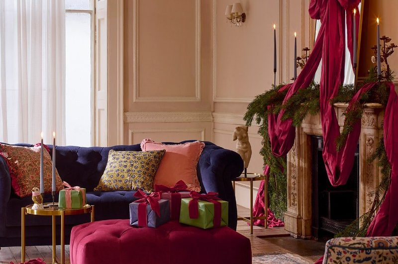 A pale pink living room with a blue sofa, red foot stool, patterned armchair and a fireplace mantel with oversized garlands and bow.