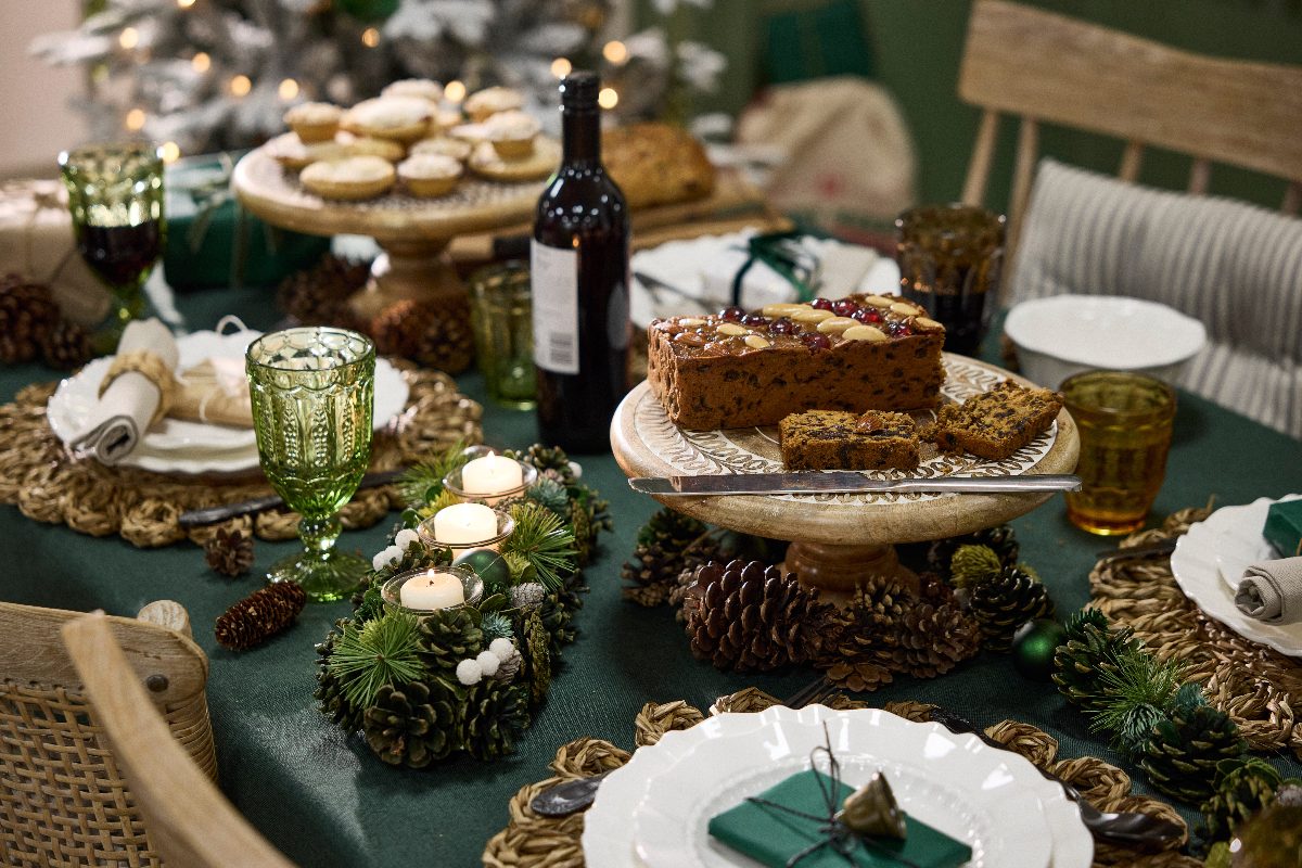 A rustic tablescape with wood cake stand, green tablecloth, rattan charger plates, and pine cone decor.