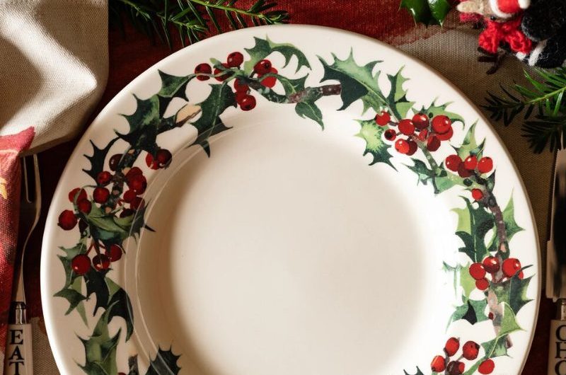 A holly berry patterned, white dinner plate on a red tablecloth.