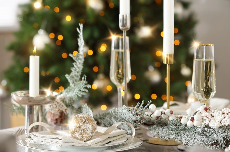 A white Christmas tablescape with frosty garland, white crockery, champagne flutes and a tree in the background.