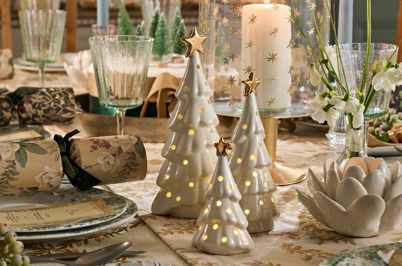 A white festive tablescape with patterned linen, ceramic trees, floral crackers and teal dinner plates.