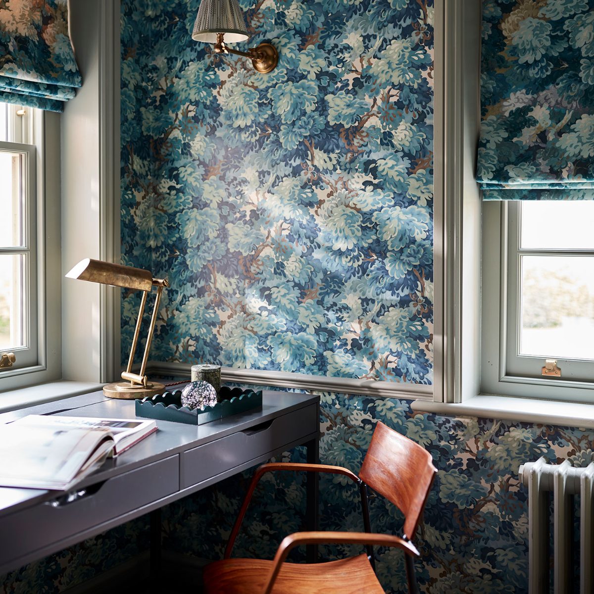 A study with floral wallcoverings, a compact desk, wood chair and windows with matching floral roller blinds.