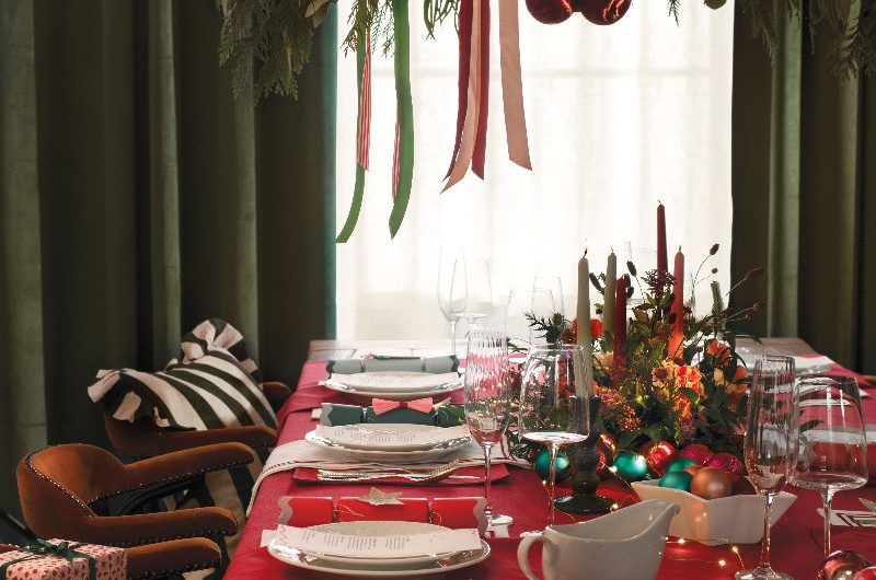 A dining room with a wood table, dressed in red tablecloth, with a festive centrepiece with candles and baubles, with white crockery, and a ceiling-mounted foliage centrepiece above it.