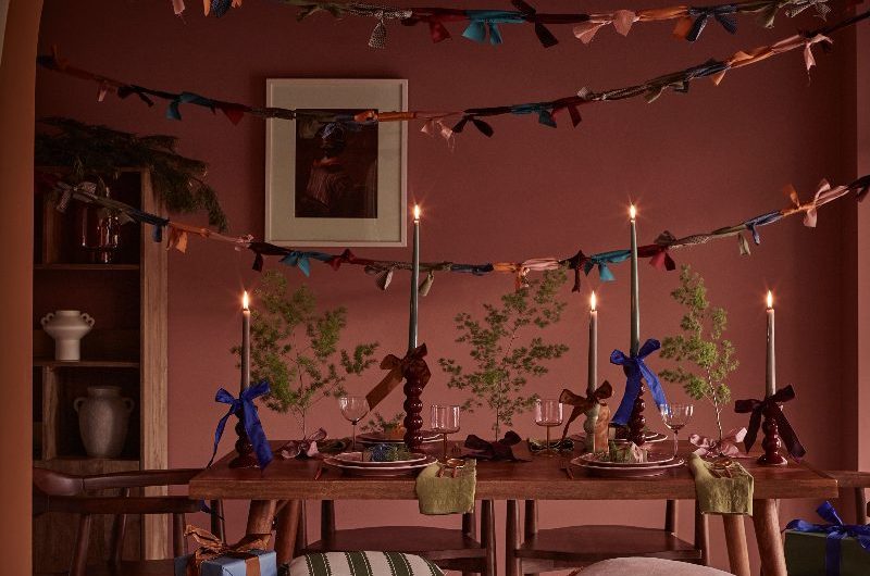 A moody Christmas dining room drenched in colour, with wood furniture, ribbon garlands, and a mix of candle sticks on the table with tied bows at the base.