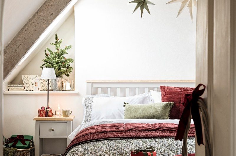 A white bedroom design decorated for Christmas with red and green furnishings, small trees, candle lanterns, star hanging decorations and stockings.