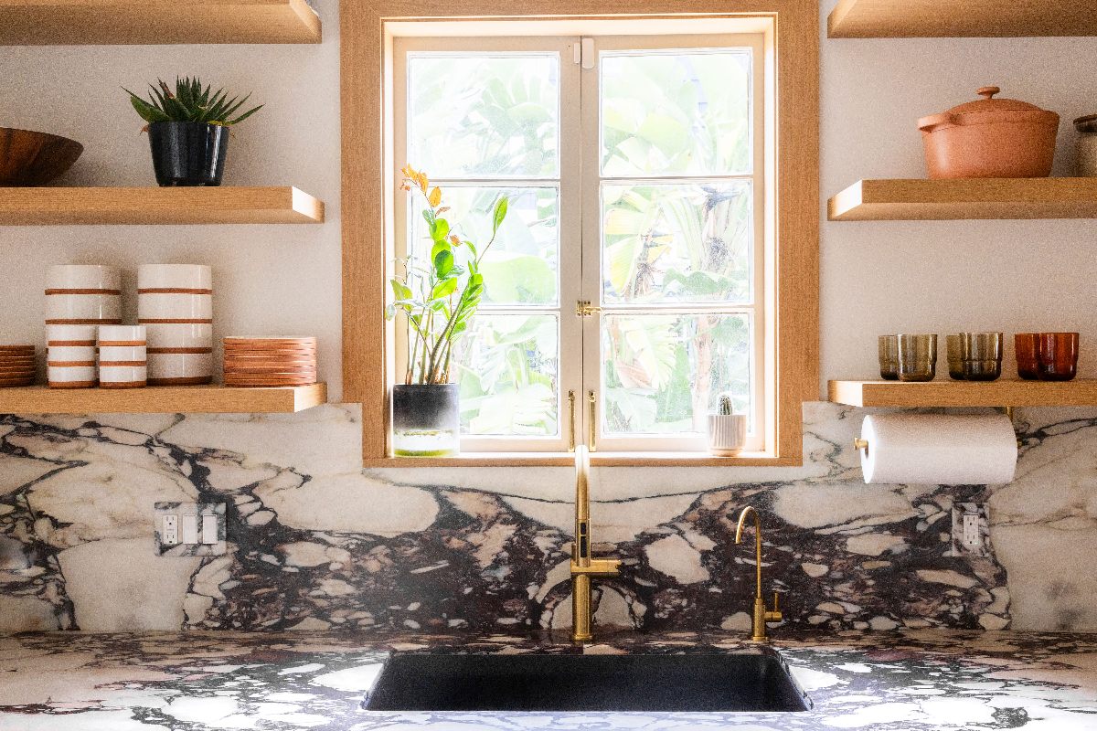A sink zone with Calacatta Viola marble, brass taps, wall-mounted oak shelves and a window.