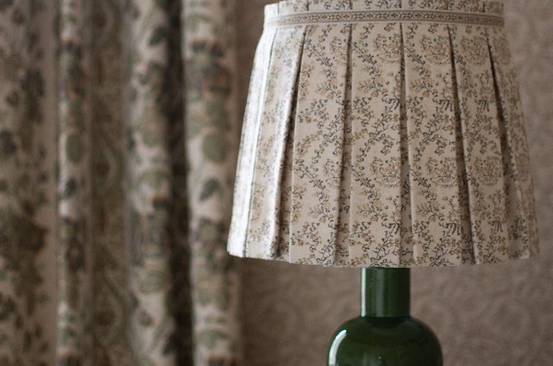 A round table with a green lamp, with floral curtains and wallpaper in the background.