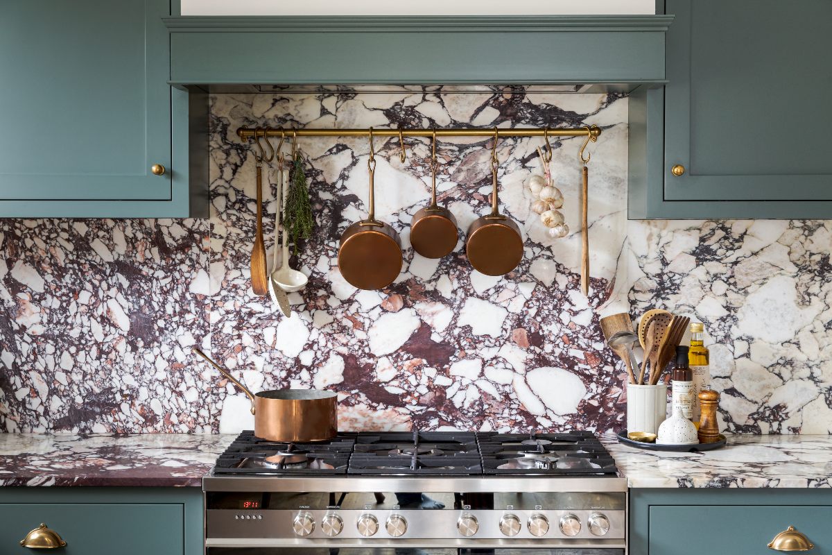 A blue kitchen design with a cooking zone, a range cooker and brass rail with hanging pots, and purply work surfaces.