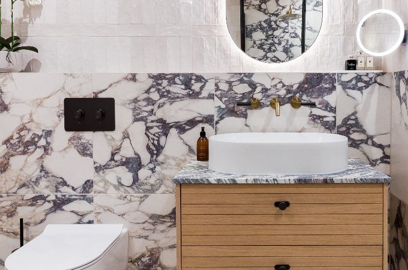 A glamorous bathroom design with glossy white metro tiles, Calacatta Viola-inspired wall tiles, elegant sanitaryware, a wood vanity unit and LED mirrors.