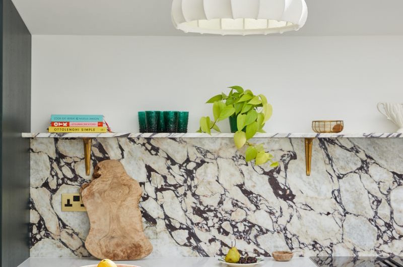 A green Shaker kitchen with a central island, coloured work surfaces, a floating shelf for displaying decor, and a white worktop on the run of cabinets with an induction hob.