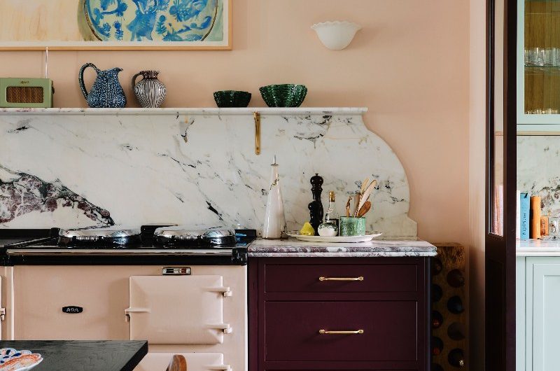 A romantic interior design with burgundy cabinetry, marble worktop and splashback which has curved detailing, a floating shelf, and pink Aga range cooker.