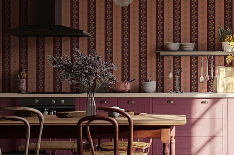 A characterful kitchen with striped and floral wallpaper, burgundy cabinetry, and central dining table with chairs.