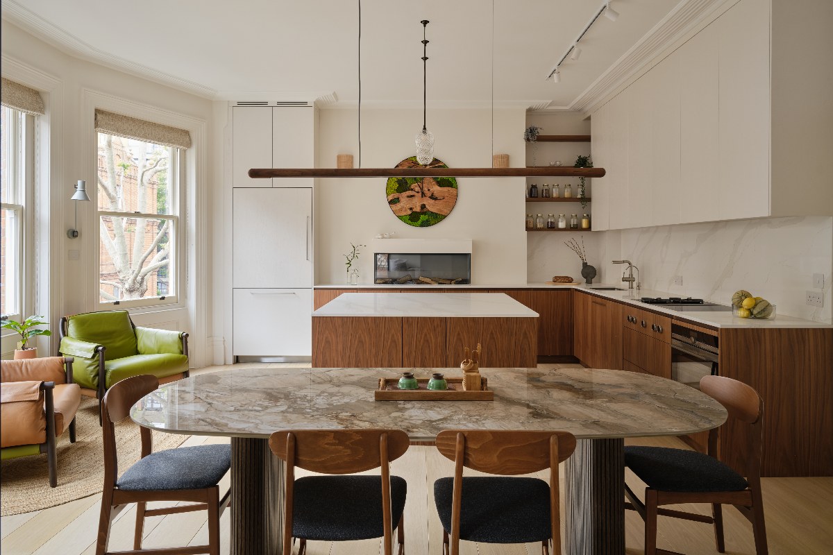Open-plan kitchen-diner with off white shades on the walls, veiny work surfaces, walnut cabinetry, leather chairs by the window and a dining area with a statement table.