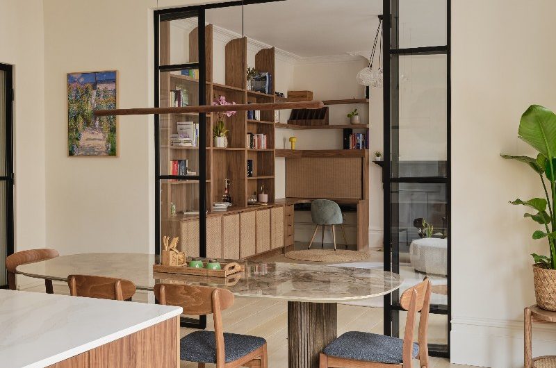 A walnut kitchen with an opening between the dining zone and library-living spaces with light flooring and off-white walls.