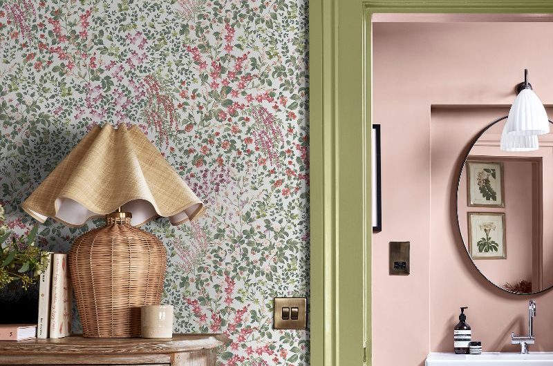 A colourful bedroom with floral wallpaper, wood chest of drawers, and a green-painted door frame leading to a pink bathroom.