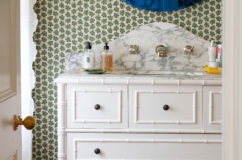 A bathroom design with green patterned wallpaper, an ornate vanity unit with marble surfaces and a blue circular mirror.