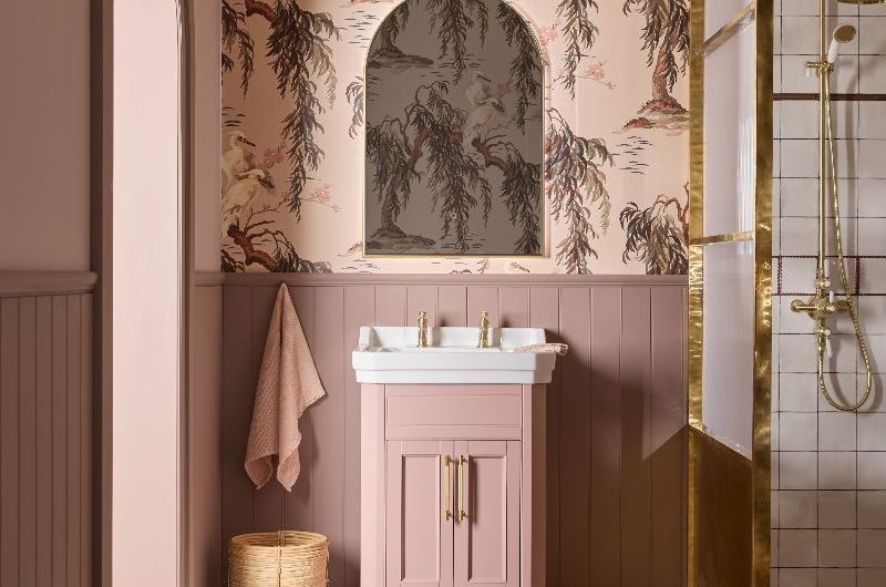 A romantic bathroom design drenched in shades of pink, with a pink vanity, wall panelling and floral wallpaper, and a walk-in shower which has a gold-framed enclosure, matching shower system and white square tiles.
